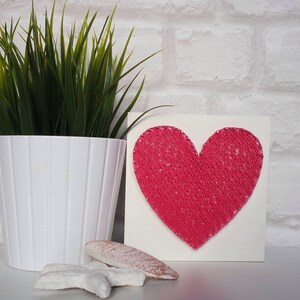 May include: A pink heart-shaped string art piece on a white background. The heart is made of pink string wrapped around nails hammered into the white board.