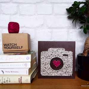 May include: A brown wooden frame with a white string art design of a camera with a pink heart in the viewfinder. The frame is sitting on a table with a stack of books and a ball of pink yarn.