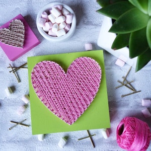 May include: A pink heart-shaped string art project on a green background. The heart is made with pink thread wrapped around nails hammered into the wood. There are also some white marshmallows and gold tacks scattered around the heart.