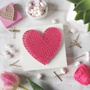 May include: A pink heart-shaped string art piece on a white background. The heart is made of pink yarn wrapped around nails hammered into the wood. There are small white marshmallows and gold tacks scattered around the heart.