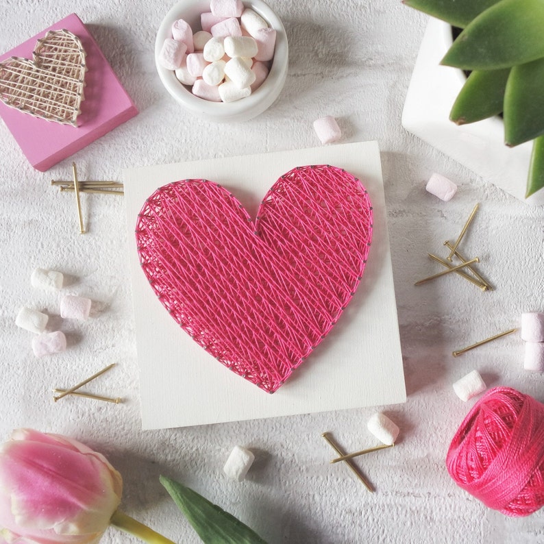 May include: A pink heart-shaped string art piece on a white background. The heart is made of pink yarn wrapped around nails hammered into the wood. There are small white marshmallows and gold tacks scattered around the heart.