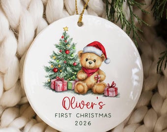 May include: A round white ceramic ornament featuring a brown teddy bear wearing a red Santa hat and scarf, sitting next to a decorated Christmas tree and gifts. The text reads Oliver's FIRST CHRISTMAS 2026 in red and black lettering.