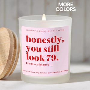 May include: A lit, frosted glass candle with a pink label that reads "honestly, you still look 79. from a distance...". The candle has a wooden lid and the text "MORE COLORS" is in the top right corner.