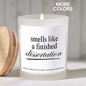 May include: A lit, frosted glass candle with a wooden lid. The candle has a white label with the text "smells like a finished dissertation." The candle is 8 oz and made of all-natural soy.