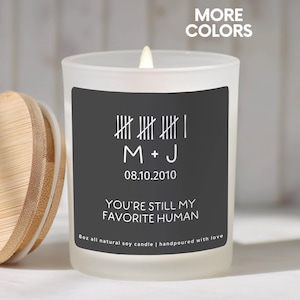 May include: A white frosted glass candle with a wooden lid. The candle has a black label with white text that reads "M + J 08.10.2010 YOU'RE STILL MY FAVORITE HUMAN". The candle is 8 oz.
