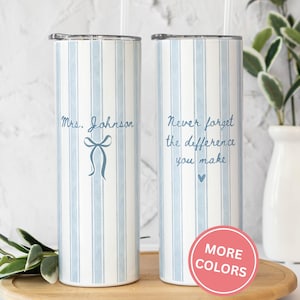 May include: Two stainless steel tumblers with blue and white vertical stripes. One tumbler has the text "Mrs. Johnson" and a bow graphic. The other tumbler has the text "Never forget the difference you make" and a heart graphic. A pink circle with "MORE COLORS" is visible.