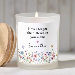 May include: A frosted glass candle with a wooden lid. The candle has a white label with the text "Never forget the difference you make" and the name "Samantha". The label features a floral design with butterflies and dragonflies.