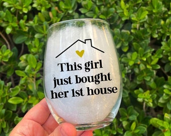 Puede incluir: Una copa de vino blanco con un diseño negro y dorado que dice "This girl just bought her 1st house".