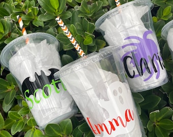May include: Three clear plastic cups with black and white striped straws. Each cup has a Halloween-themed design and a name written on it: "Scott", "Emma", and "Chris".