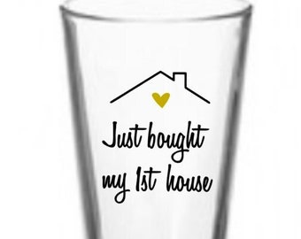 Puede incluir: Un vaso de vidrio transparente con un diseño negro y dorado que dice "Just bought my 1st house" con un pequeño corazón sobre la palabra "bought".