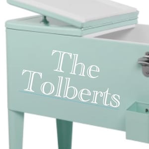 May include: A mint green cooler with a white interior and a hinged lid. The cooler has a bottle opener, a small storage compartment, and the text "The Tolberts" on the side. It is elevated on four matching legs.