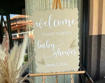 May include: A baby shower sign with the text "Welcome to Baby Tolbert's Baby Shower January 2, 2022" on a wooden easel. A vase with dried pampas grass sits on a table with a pink napkin and a glass of water.