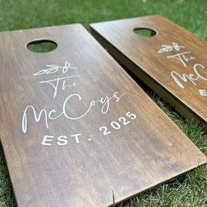 May include: Two wooden cornhole boards with a dark stain. Each board has a circular hole and the text "The McCoys EST. 2025" in white script. The boards are on a green grassy surface.