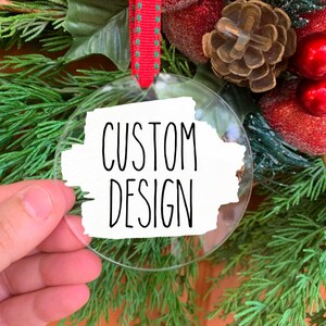 May include: A clear glass Christmas ornament with a white background and black text that reads "CUSTOM DESIGN".