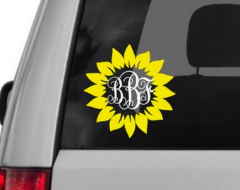 May include: A yellow sunflower decal with a white monogram of the letters "BBS" in the center. The decal is on a gray car window.
