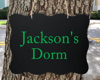 Puede incluir: Un letrero negro de pizarra con un borde ondulado cuelga de un árbol. El letrero dice "Jackson's Dorm" en letras verdes.