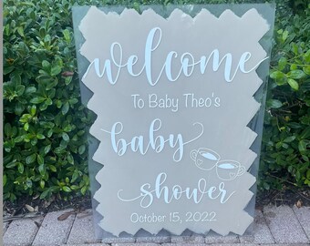 May include: A white welcome sign with the text "Welcome to Baby Theo's Baby Shower October 15, 2022" and two small teacups with hearts on them.