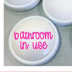 May include: White, round bathroom sign with the words "bathroom in use" in bright pink script. The sign has a slightly raised edge and is likely made of plastic or ceramic. Several signs are visible.