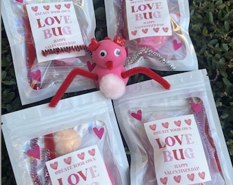May include: Four clear plastic bags with craft kits for making Love Bugs. Each bag has a label saying "Create Your Own Love Bug Happy Valentine's Day!" and contains pipe cleaners, pom-poms, and other craft materials. A pink Love Bug is in the center.
