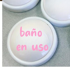 May include: White, round bathroom sign with a raised edge. The sign has the words "baño en uso" in pink script. The sign is likely used to indicate if a bathroom is occupied.