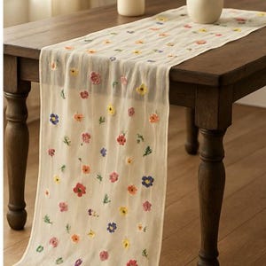 May include: A cream-colored table runner with embroidered flowers in shades of red, pink, yellow, and blue. The runner is draped over a dark wooden table, adding a touch of floral elegance to the dining area.