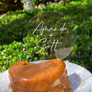 May include: A clear round acrylic sign with the text "Amanda + Scott" written in white cursive script. The sign is sitting on top of a small cake with caramel icing.