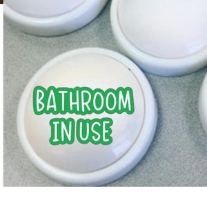May include: White, round bathroom sign with the text "BATHROOM IN USE" in green. The sign is slightly raised and has a rounded edge. Several similar signs are visible in the image.
