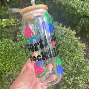 May include: A clear glass reusable water bottle with a bamboo lid and a straw. The bottle has a watermelon, blueberry, and strawberry design with the text "cortisol cocktail" in black.
