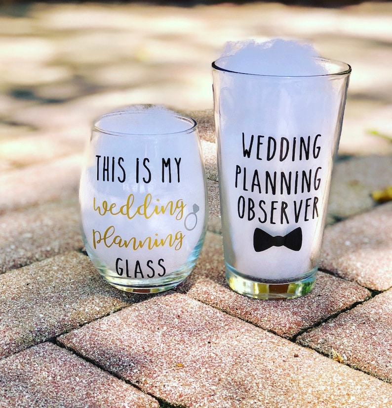 May include: Two glasses with text on them. The smaller glass is a wine glass with the text "This is my wedding planning glass". The larger glass is a pint glass with the text "Wedding planning observer" and a black bow tie.