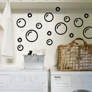 May include: A laundry room scene featuring two white washing machines with the brand name "Miele". A woven basket and a metal container sit atop the machines. Black bubble decals adorn the wall, and two white towels hang from a rack.