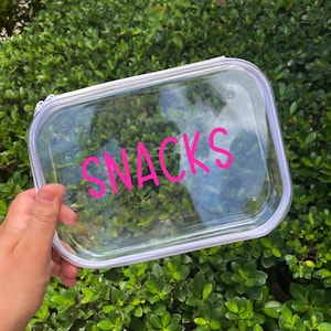 May include: A clear rectangular container with a white zipper and the word "SNACKS" in bright pink lettering. The container is held against a backdrop of green foliage.