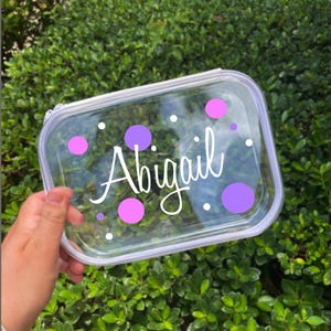 May include: Clear plastic zippered pouch with a white background and purple polka dots. The name "Abigail" is written in white cursive lettering.