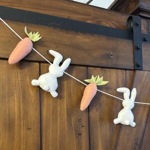 May include: A white felt bunny and orange felt carrot garland with green felt leaves. The garland is strung on a white string and is hanging on a wooden door.