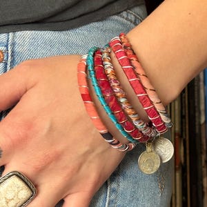 May include: A stack of colorful fabric bracelets with silver coin charms. The bracelets are made of red, pink, and turquoise fabric and are wrapped around a person's wrist.