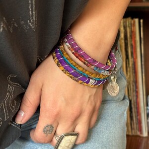 May include: A set of five colorful fabric bracelets with a silver charm. The bracelets are layered on a person's wrist and feature purple, yellow, and blue colors.