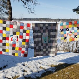 May include: Three colourful baby quilts with a patchwork design are hanging on a clothesline. The quilts feature a dog applique with the names "Max", "Doodle", and "Peaches" embroidered on them.