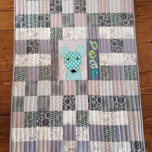 May include: A grey and white patchwork baby quilt with a green and white dog applique in the centre. The quilt is quilted with a straight stitch pattern and has the name "Beau" embroidered in the corner.
