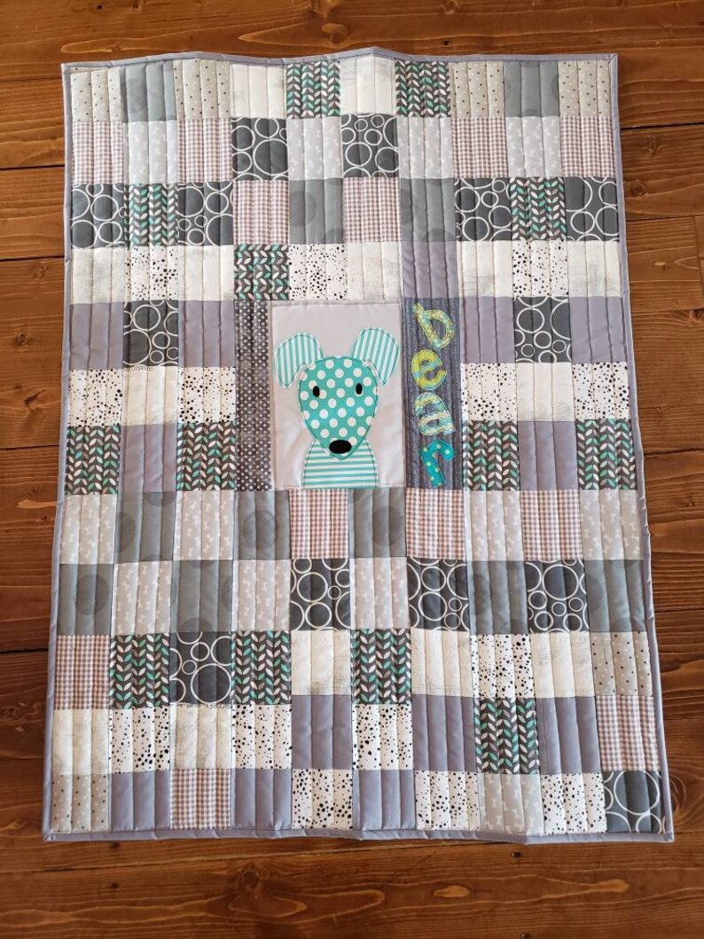 May include: A grey and white patchwork baby quilt with a green and white dog applique in the centre. The quilt is quilted with a straight stitch pattern and has the name "Beau" embroidered in the corner.