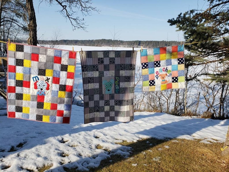 May include: Three colourful baby quilts with a patchwork design are hanging on a clothesline. The quilts feature a dog applique with the names "Max", "Doodle", and "Peaches" embroidered on them.