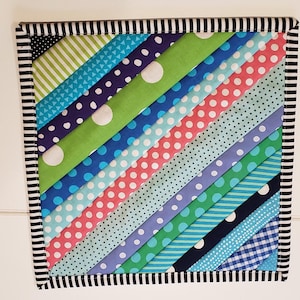 May include: A colorful patchwork pot holder with a diagonal pattern of green, blue, pink, and white fabric strips. The pot holder is edged with a black and white striped border.