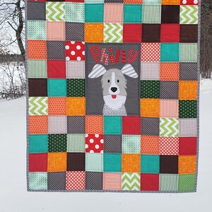 May include: A colourful patchwork quilt with a grey square in the centre featuring a cartoon dog with the name "Phineas" embroidered in red. The quilt is made up of squares of various colours and patterns, including red, orange, yellow, green, blue, and brown.