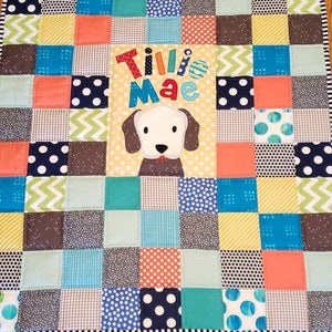 May include: A patchwork baby quilt with a variety of colourful squares, including blue, green, yellow, and orange. The quilt features a white dog applique with the name "Tillie Mae" embroidered in colourful letters.