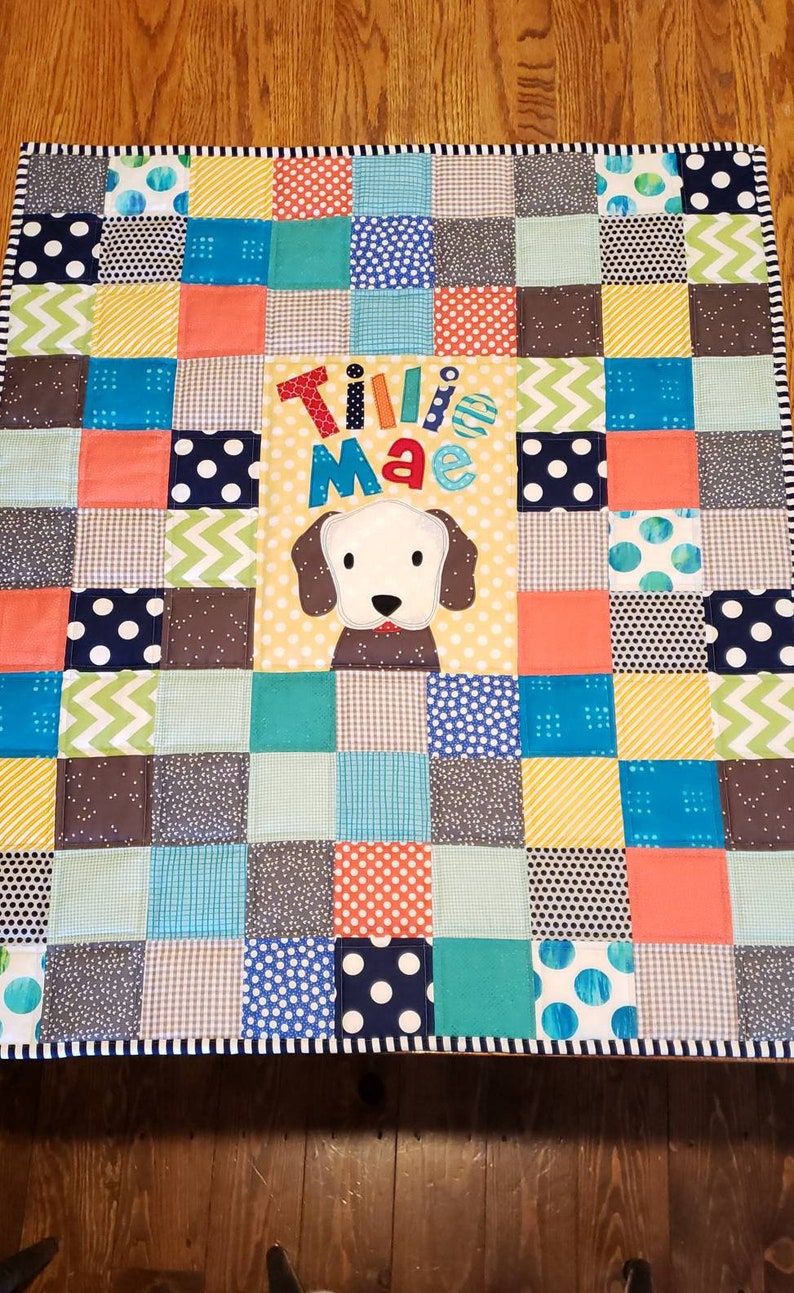 May include: A patchwork baby quilt with a variety of colourful squares, including blue, green, yellow, and orange. The quilt features a white dog applique with the name "Tillie Mae" embroidered in colourful letters.