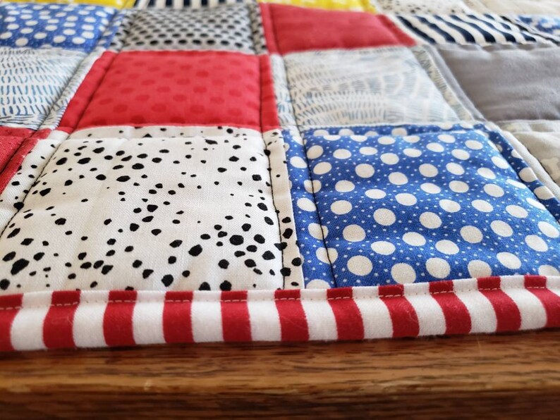 May include: A patchwork quilt featuring a red, white and blue colour scheme. The quilt is made from different fabrics with various patterns, including polka dots, stripes and geometric designs. The quilt has a red and white striped border.