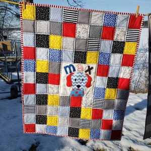 May include: A red, white and blue patchwork baby quilt with a white square in the centre featuring a blue and grey dog with the name "Max" embroidered on it.