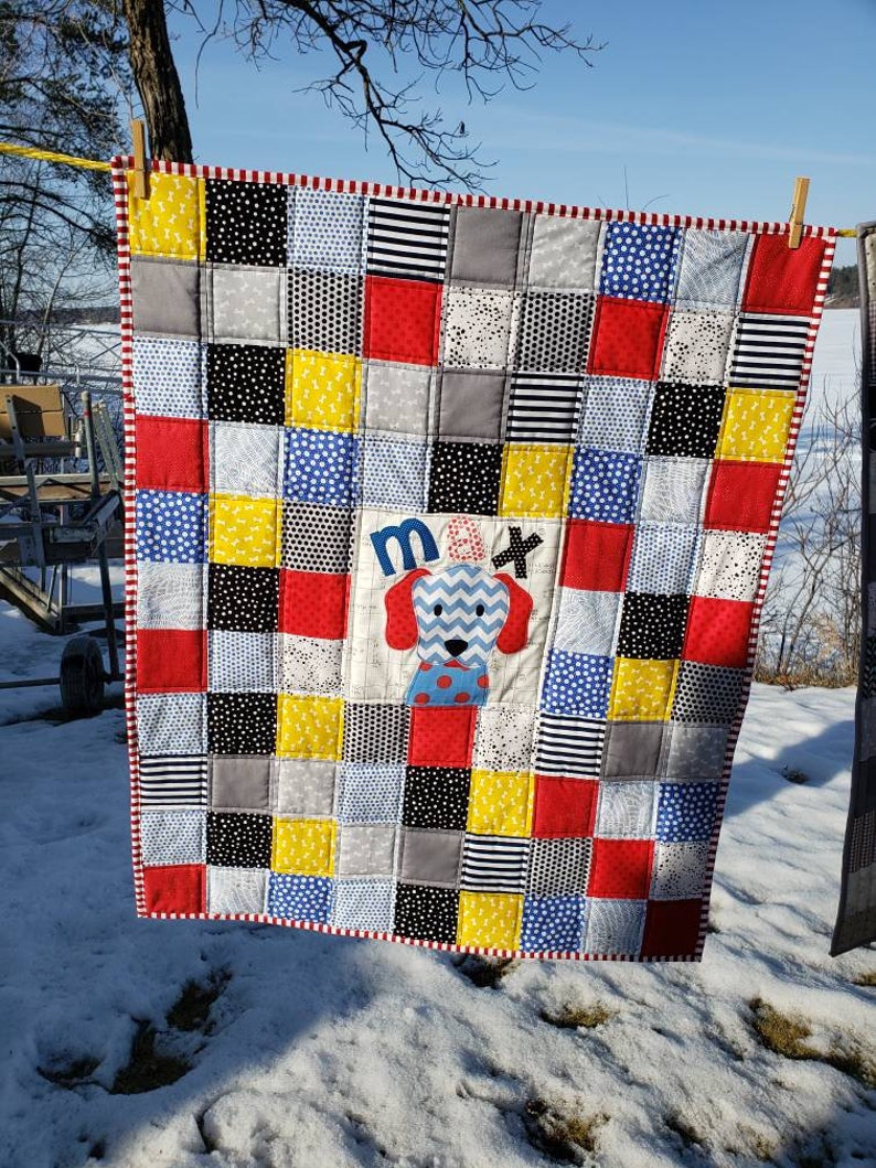 May include: A red, white and blue patchwork baby quilt with a white square in the centre featuring a blue and grey dog with the name "Max" embroidered on it.