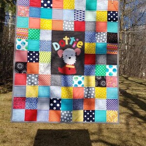 May include: A colourful patchwork quilt featuring a variety of fabrics in red, blue, yellow, green and black. The quilt has a central panel with the name "Dottie" and a white dog applique.