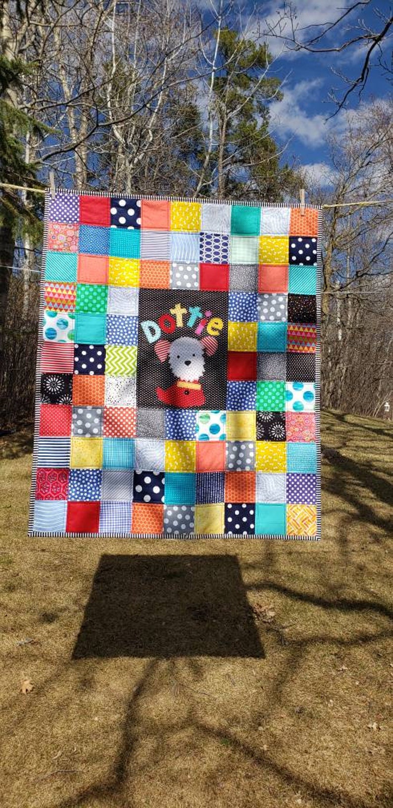 May include: A colourful patchwork quilt featuring a variety of fabrics in red, blue, yellow, green and black. The quilt has a central panel with the name "Dottie" and a white dog applique.