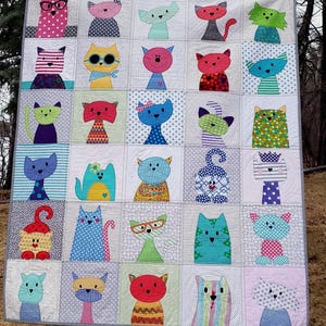 May include: A handmade quilt featuring a patchwork design of whimsical cat faces. Each square showcases a unique cat with different colors, patterns, and accessories like glasses and bows. The quilt is displayed outdoors, hanging from a clothesline.