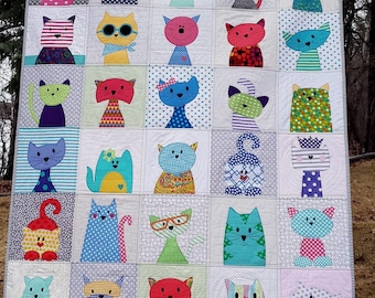 May include: A handmade quilt featuring a patchwork design of whimsical cat faces. Each square showcases a unique cat with different colors, patterns, and accessories like glasses and bows. The quilt is displayed outdoors, hanging from a clothesline.